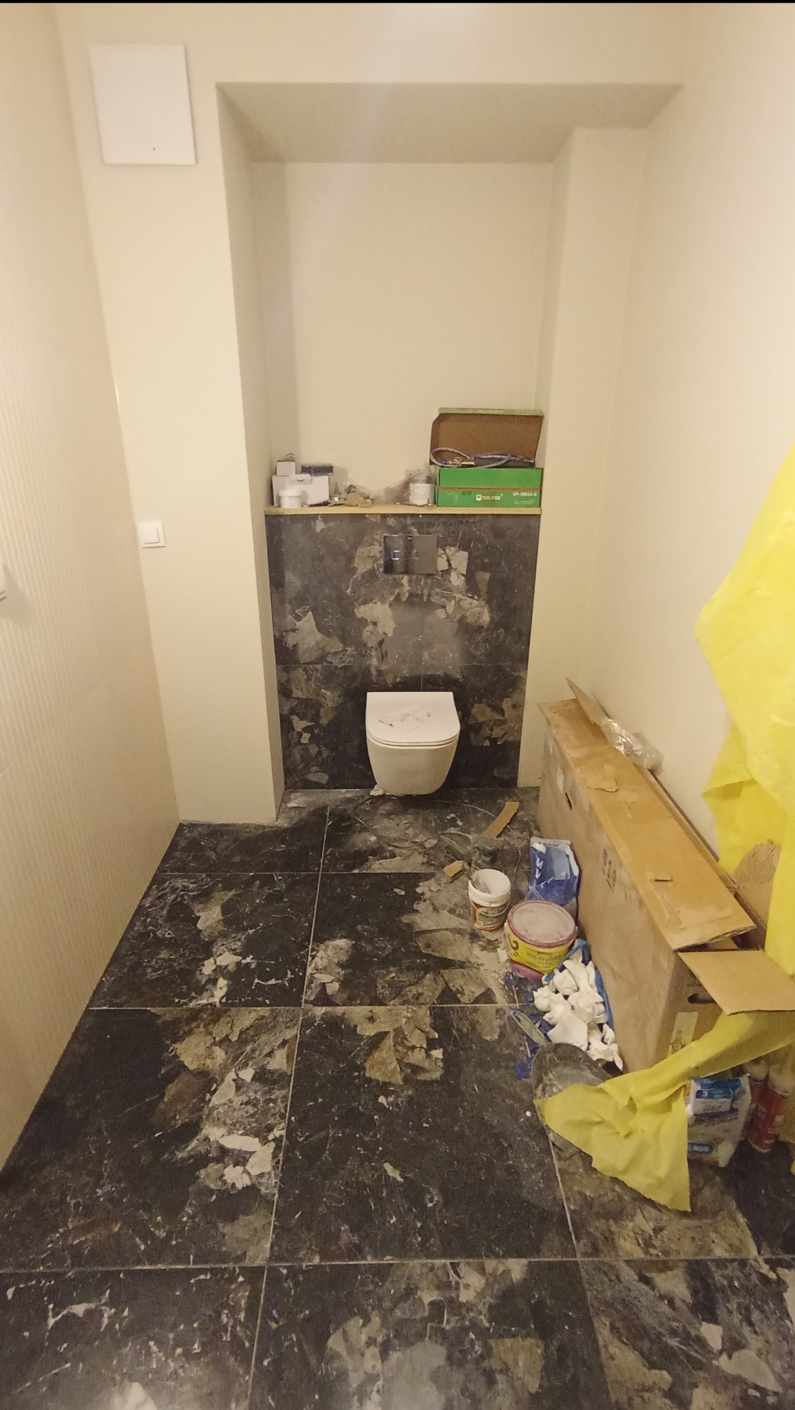 Łazienka w trakcie prac: częściowo położone ciemne płytki imitujące marmur na podłodze i ścianie, wisząca toaleta, widoczne narzędzia i materiały budowlane, żółta folia ochronna.