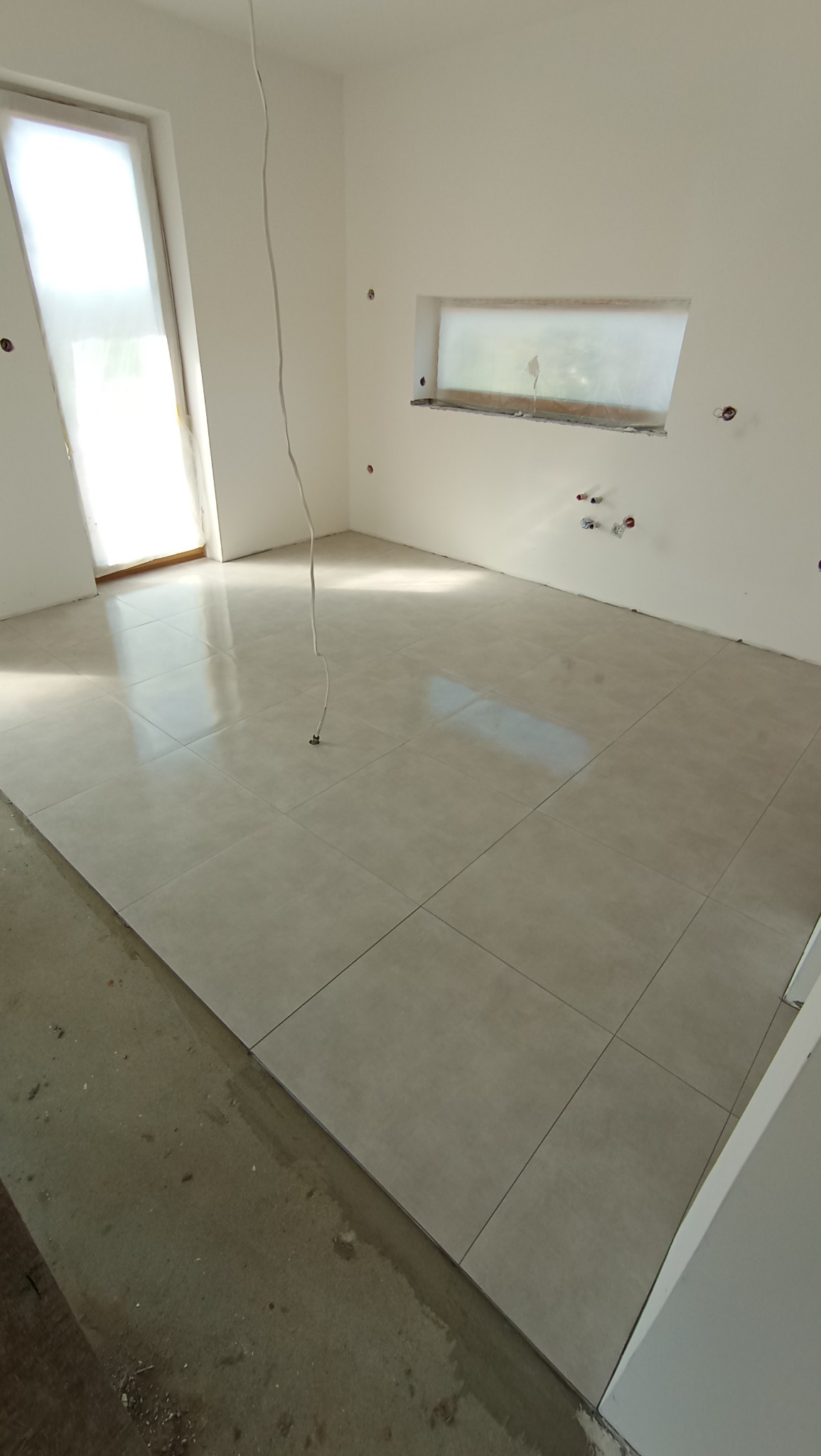 Ułożone, beżowe płytki podłogowe w jasnym pomieszczeniu z oknem i instalacją elektryczną w Kamienicy Szlacheckiej. Widoczne fugi i fragment surowej podłogi.