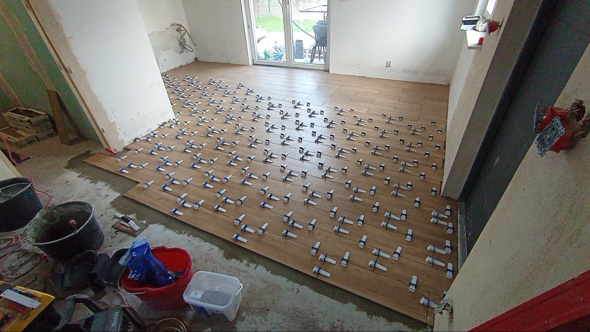 Układanie drewnopodobnych płytek podłogowych z użyciem systemu poziomowania. Widoczne narzędzia i materiały budowlane. Praca w toku, jasne wnętrze z oknem balkonowym.