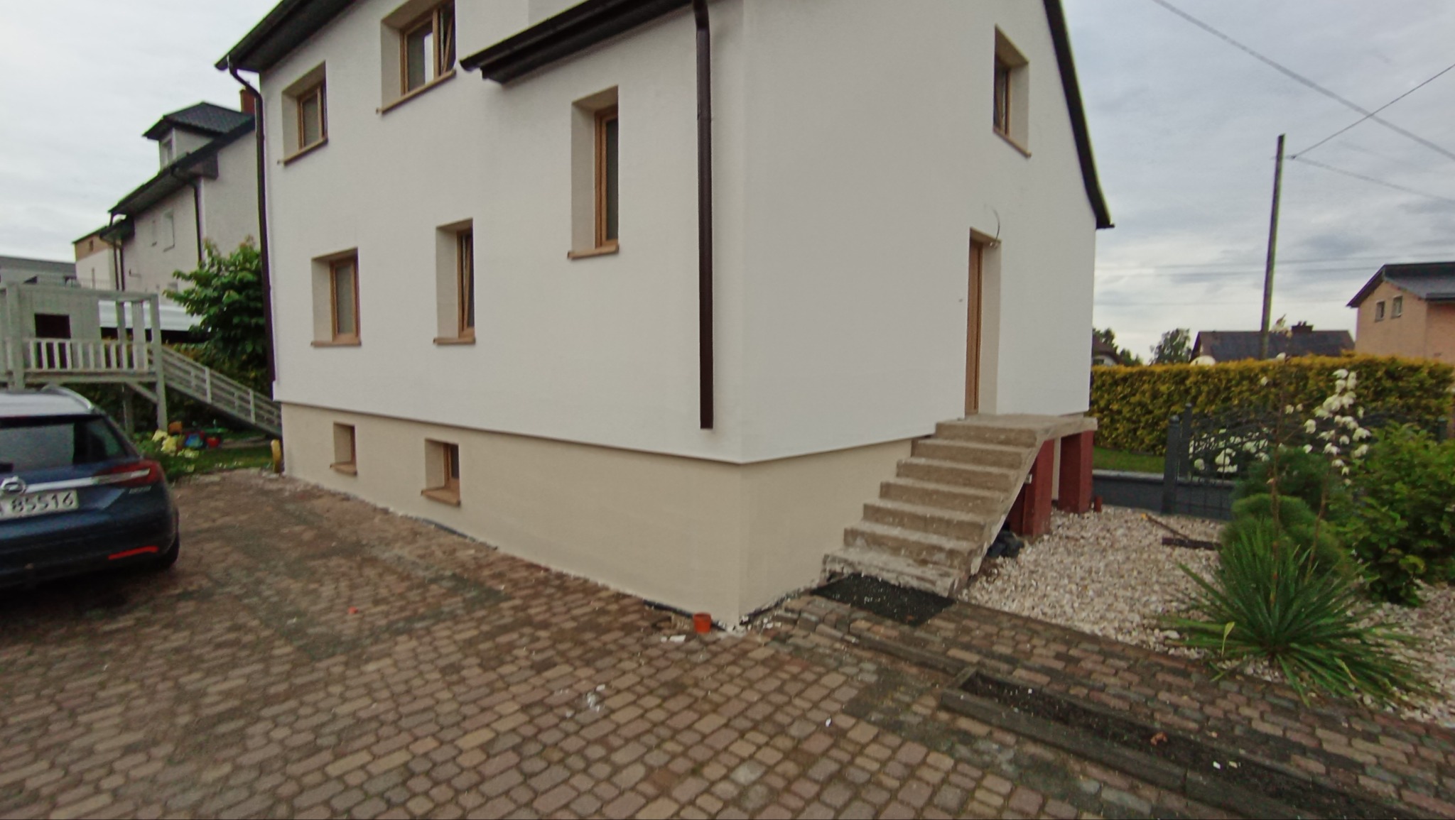 Elewacja dwukondygnacyjnego domu po remoncie, z widocznym ociepleniem fundamentów i nowym tynkiem. Na pierwszym planie kostka brukowa i zaparkowany samochód.