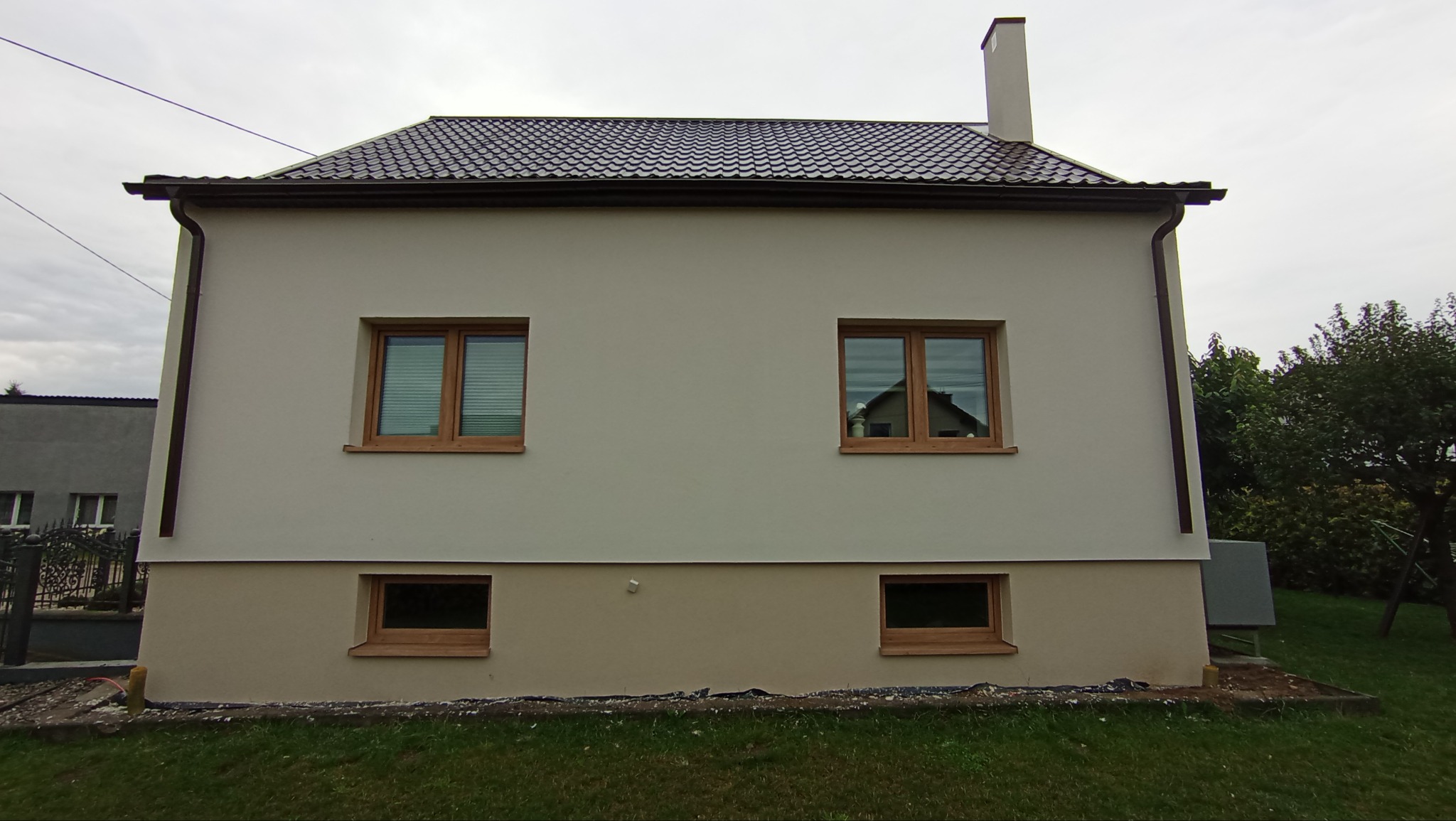 Elewacja domu jednorodzinnego po remoncie, z widocznymi oknami z drewnianymi ramami i świeżo pomalowaną fasadą w odcieniach beżu i brązu, na tle pochmurnego nieba.