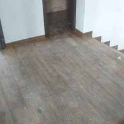 VadMet FIRMA OGÓLNOBUDOWLANA VADYM METALINSKYI - Ułożona podłoga z płytek imitujących drewno, widok na wejście do łazienki wykończonej podobnymi płytkami, białe ściany i listwy przypodłogowe.