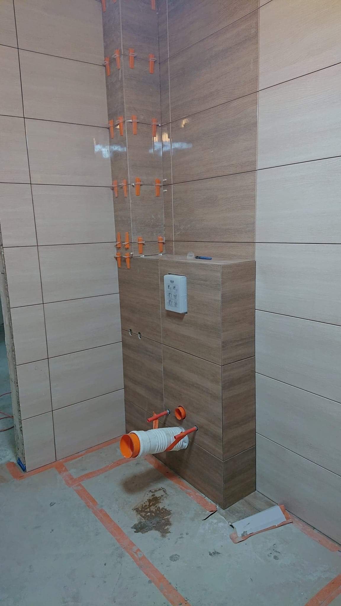 Montaż płytek imitujących drewno w łazience, widoczne pomarańczowe klipsy poziomujące, instalacja podtynkowego systemu WC w trakcie prac, zarys podziału stref na podłodze.