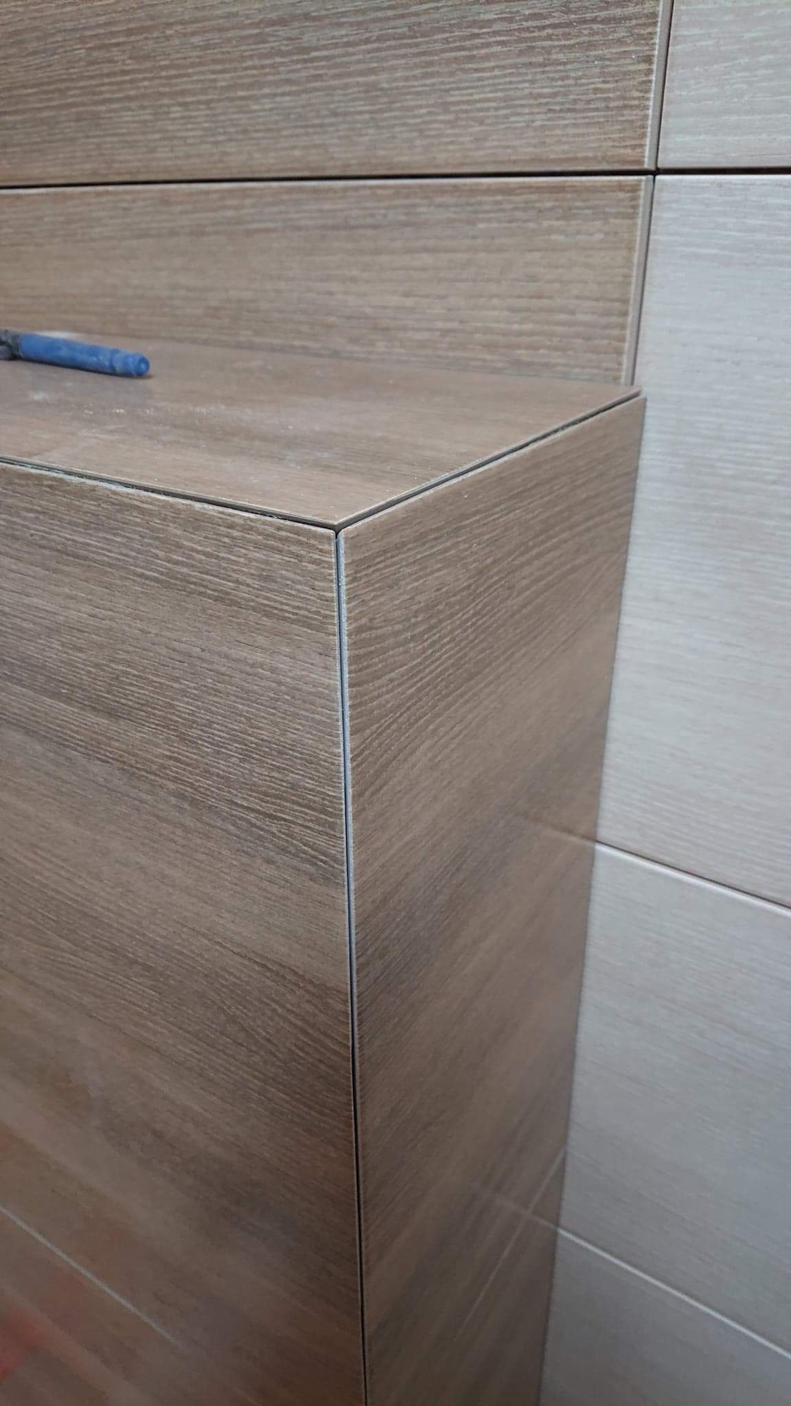 Fragment obudowy z płytek imitujących drewno, z widocznym niebieskim narzędziem glazurniczym. Widoczne fugi i precyzyjne wykończenie narożnika.