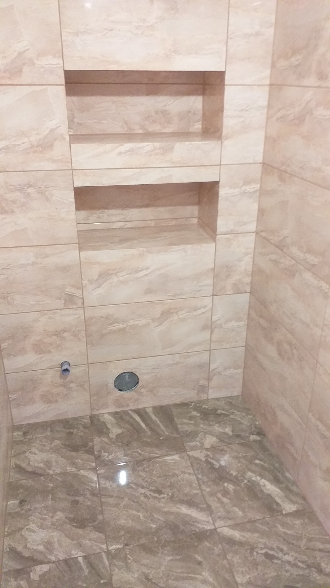 Wykończona łazienka z beżowymi płytkami imitującymi marmur, w ścianie wnęki półkowe, widoczny otwór na rurę i odpływ w podłodze.