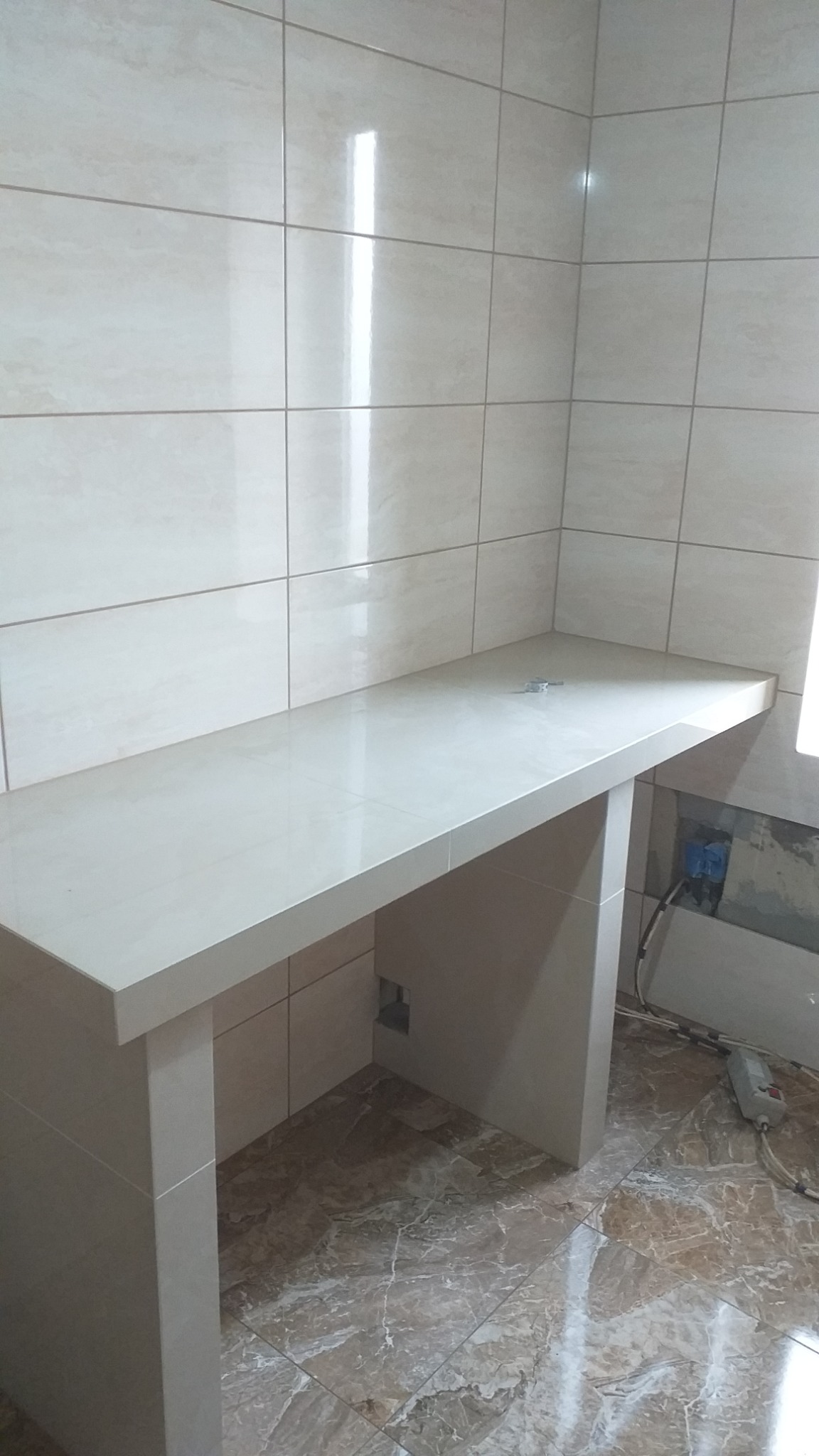 Wykończona półka pod umywalkę w łazience, wyłożona beżowymi płytkami ceramicznymi, dopasowanymi kolorystycznie do płytek na ścianach i podłodze, z widocznym gniazdkiem elektrycznym w ścianie.