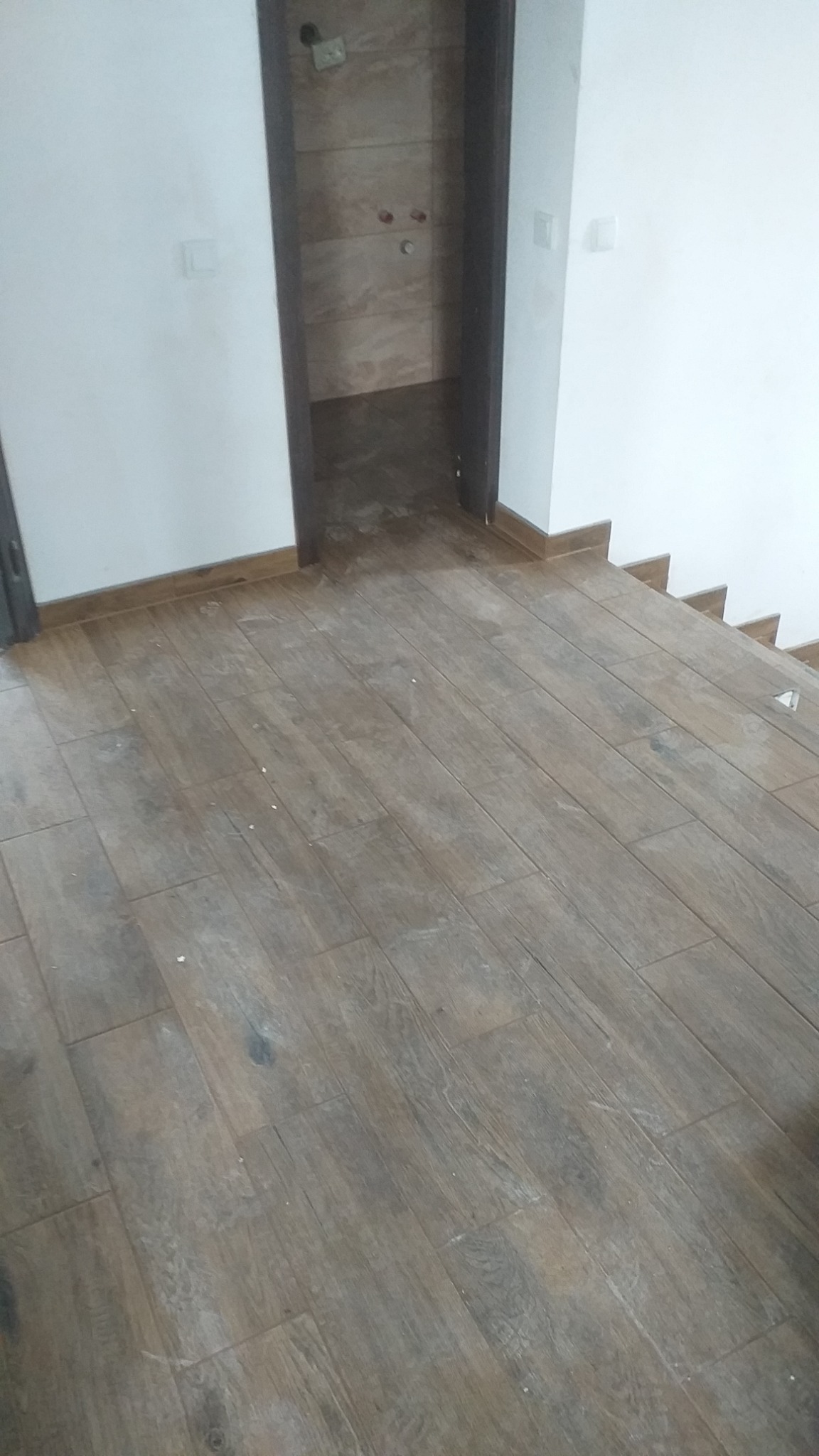 Ułożona podłoga z płytek imitujących drewno, widok na wejście do łazienki wykończonej podobnymi płytkami, białe ściany i listwy przypodłogowe.