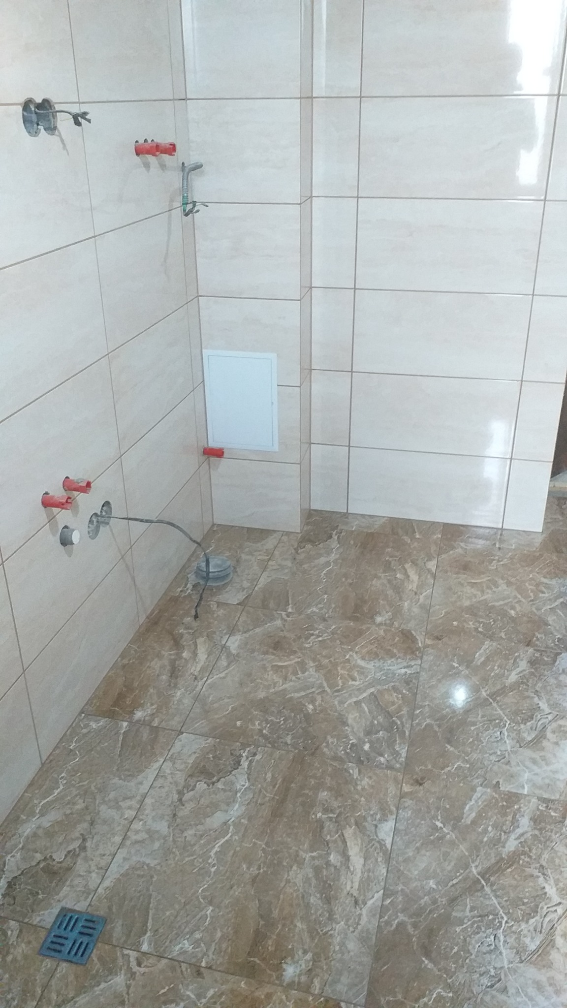 Świeżo wykafelkowana łazienka z beżowymi płytkami na ścianach i marmurkową podłogą, widoczne niedokończone przyłącza wodne i odpływ podłogowy.