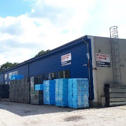 POL-MATERIAŁY BUDOWLANE SPÓŁKA Z OGRANICZONĄ ODPOWIEDZIALNOŚCIĄ - Skład budowlany z zapasem styropianu i wełny mineralnej na zewnątrz. Budynek w kolorze niebieskim z szarymi elementami. Słoneczny dzień, błękitne niebo z chmurami.