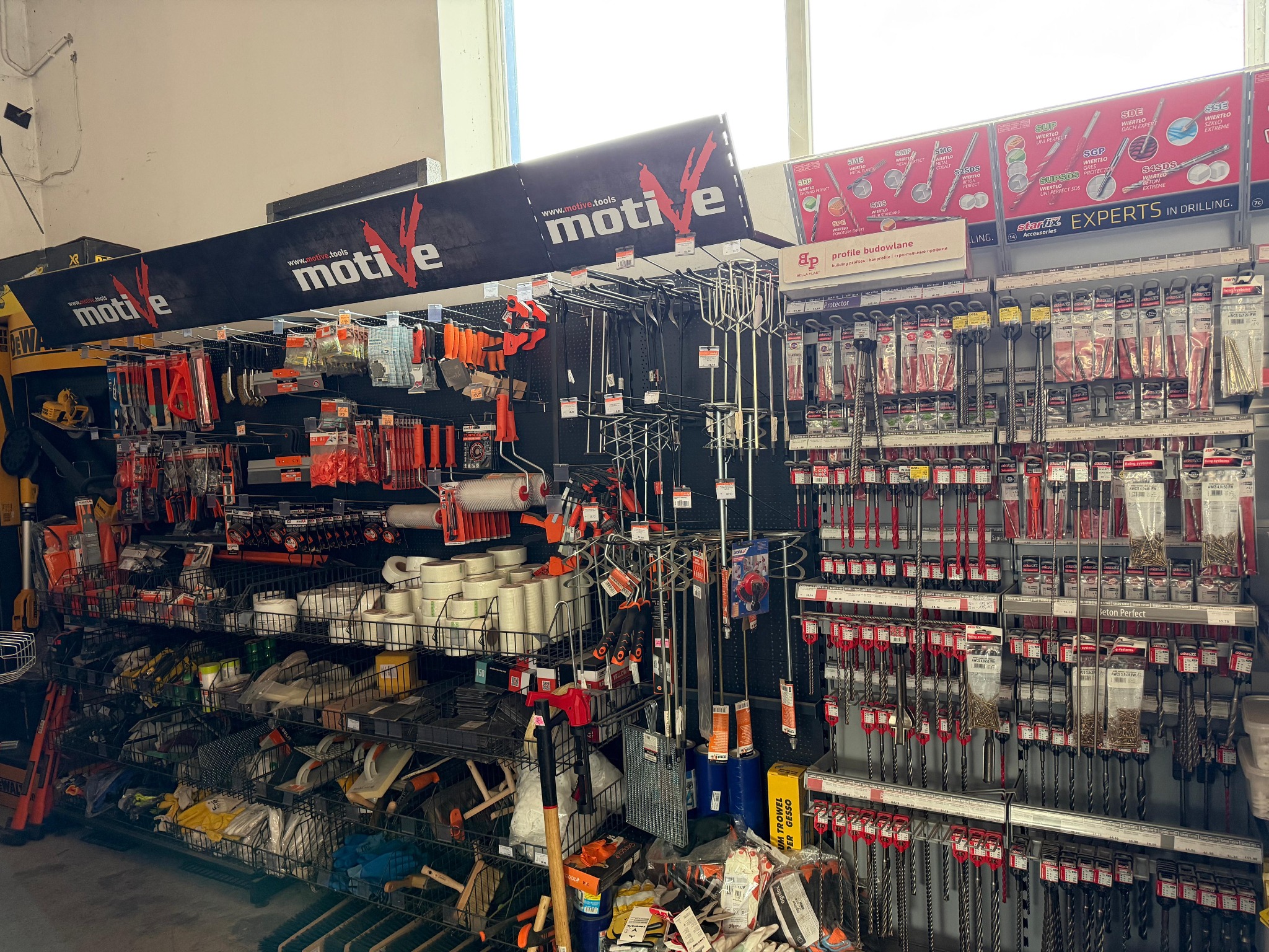 Półki w składzie budowlanym z narzędziami i akcesoriami marki Motive, wiertła, mieszadła, wałki, taśmy. Ekspozycja produktów w sklepie.