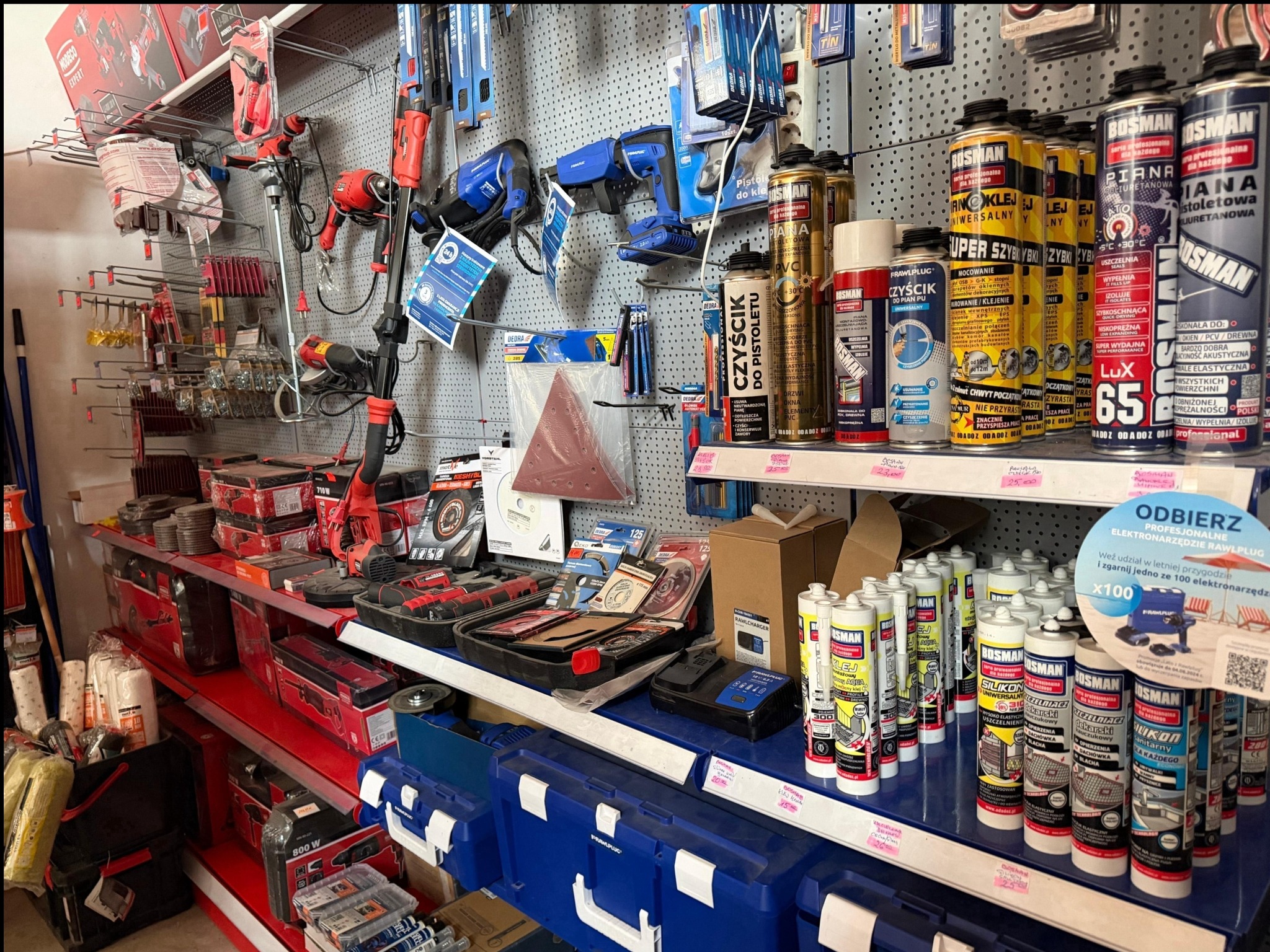 Półki w sklepie budowlanym: narzędzia, pianki montażowe, silikony, wiertarki, szlifierki, kleje, pistolety do piany, skrzynki narzędziowe, materiały ścierne.