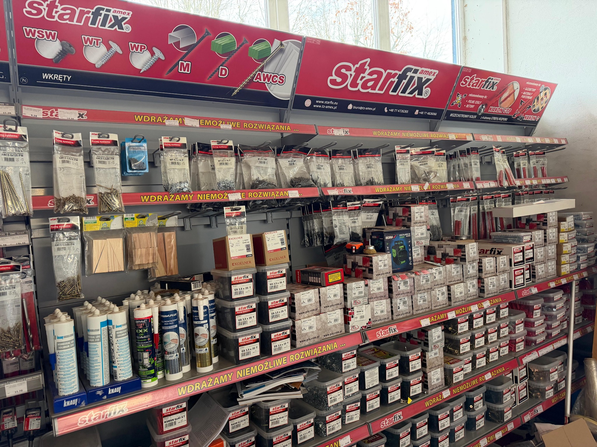 Półki w składzie budowlanym wypełnione opakowaniami wkrętów, kołków i pian montażowych marki Starfix. Ekspozycja materiałów budowlanych w sklepie.