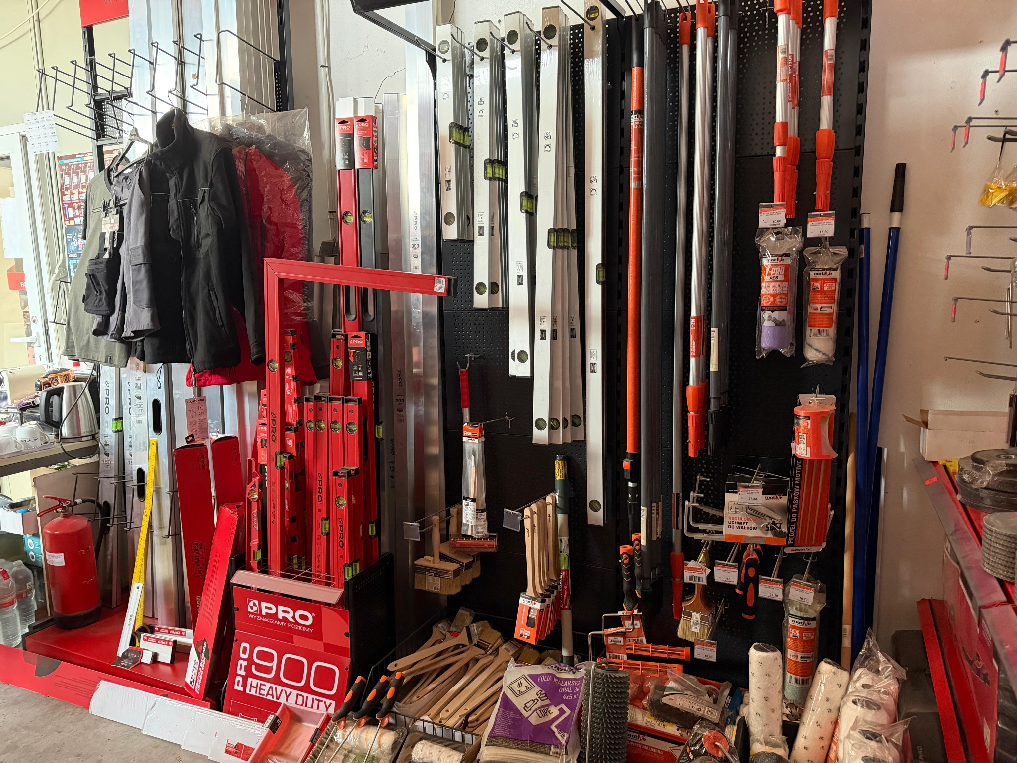 Asortyment narzędzi budowlanych: poziomice, pędzle malarskie, wałki i odzież robocza w sklepie. Czerwone akcenty dominują w ekspozycji.