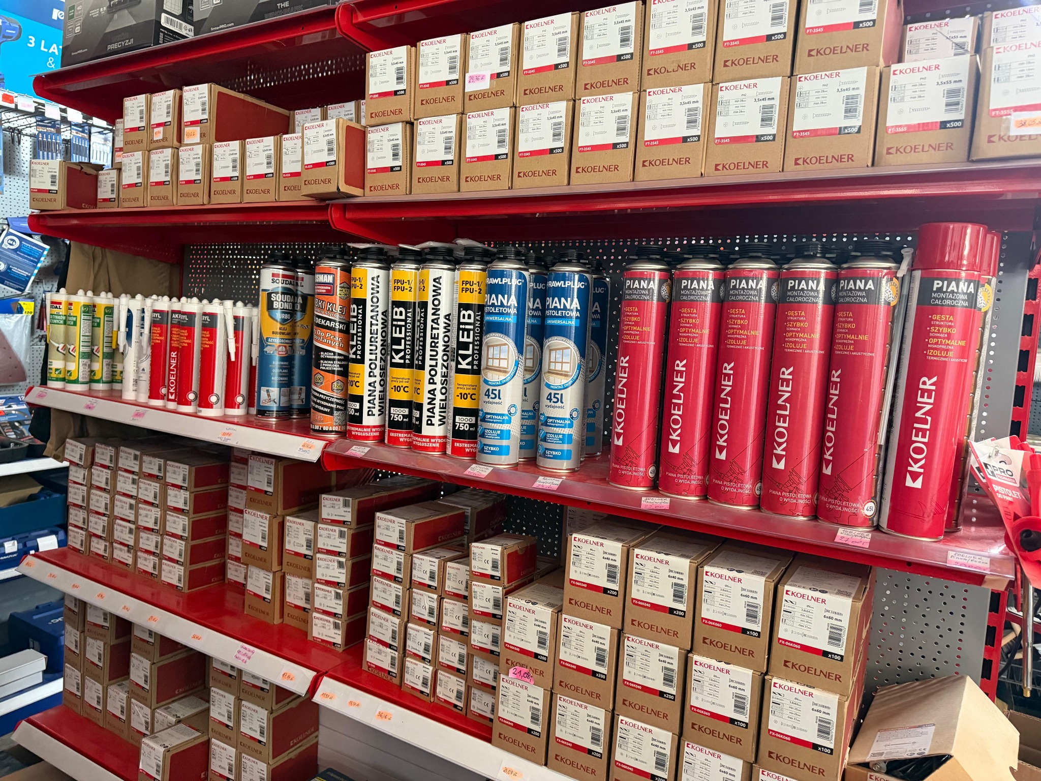Półki w sklepie budowlanym: piany montażowe Kleib i Koelner, kotwy i wkręty w kartonach. Asortyment dobrze oświetlony, widok z bliska.