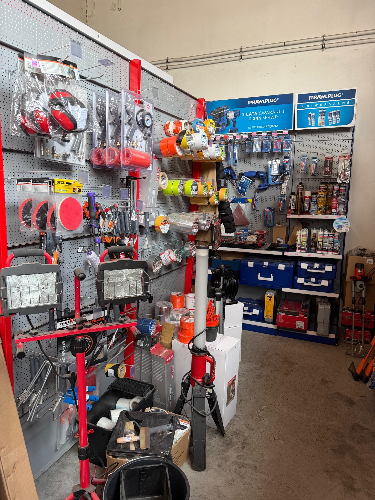 Ekspozycja narzędzi i akcesoriów budowlanych w sklepie: szlifierki, wiertarki, taśmy, pędzle i inne. Widoczne oświetlenie warsztatowe i reklamy producentów.