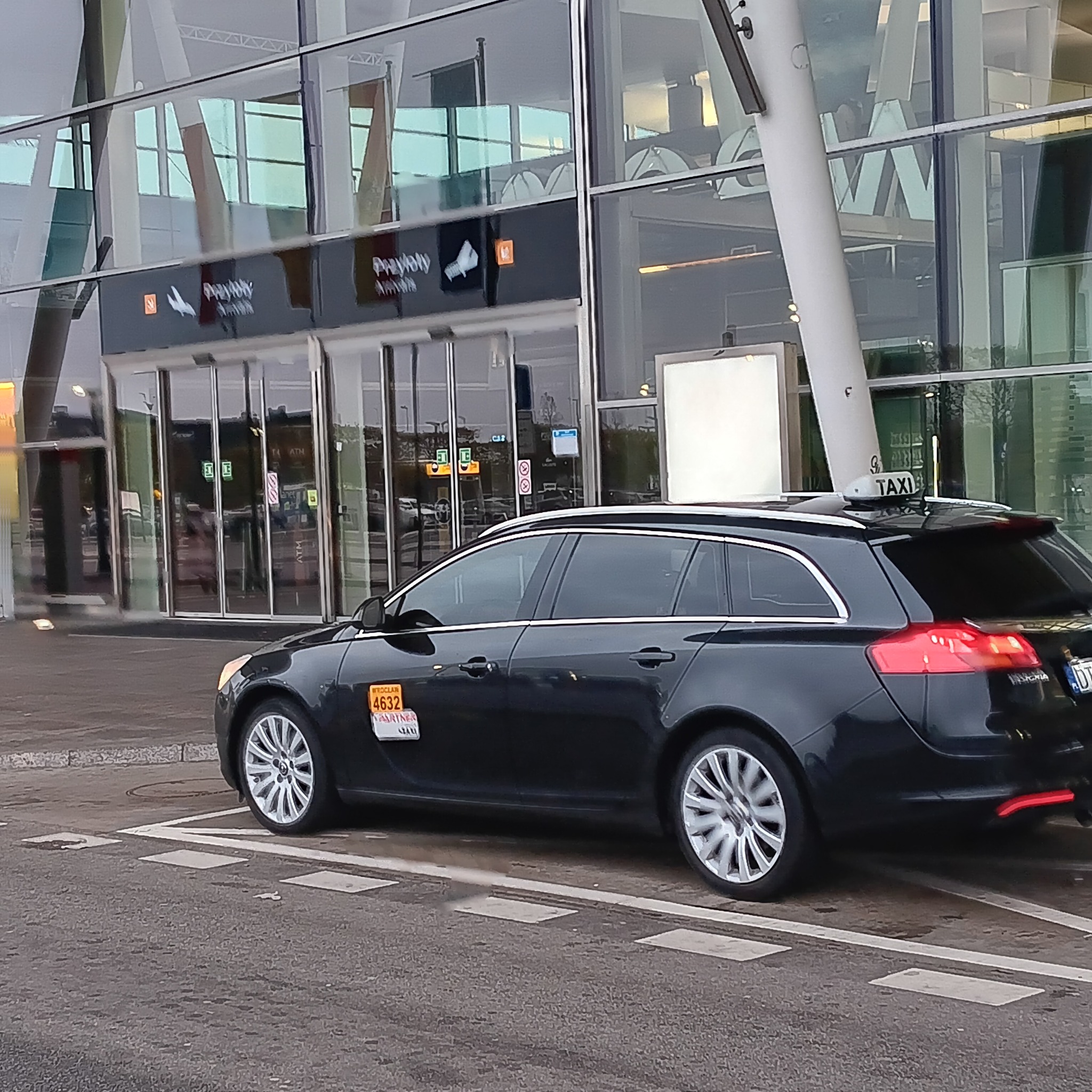 Czarny samochód typu kombi z napisem TAXI na dachu, zaparkowany przed nowoczesnym budynkiem lotniska z przeszkloną fasadą i automatycznymi drzwiami.