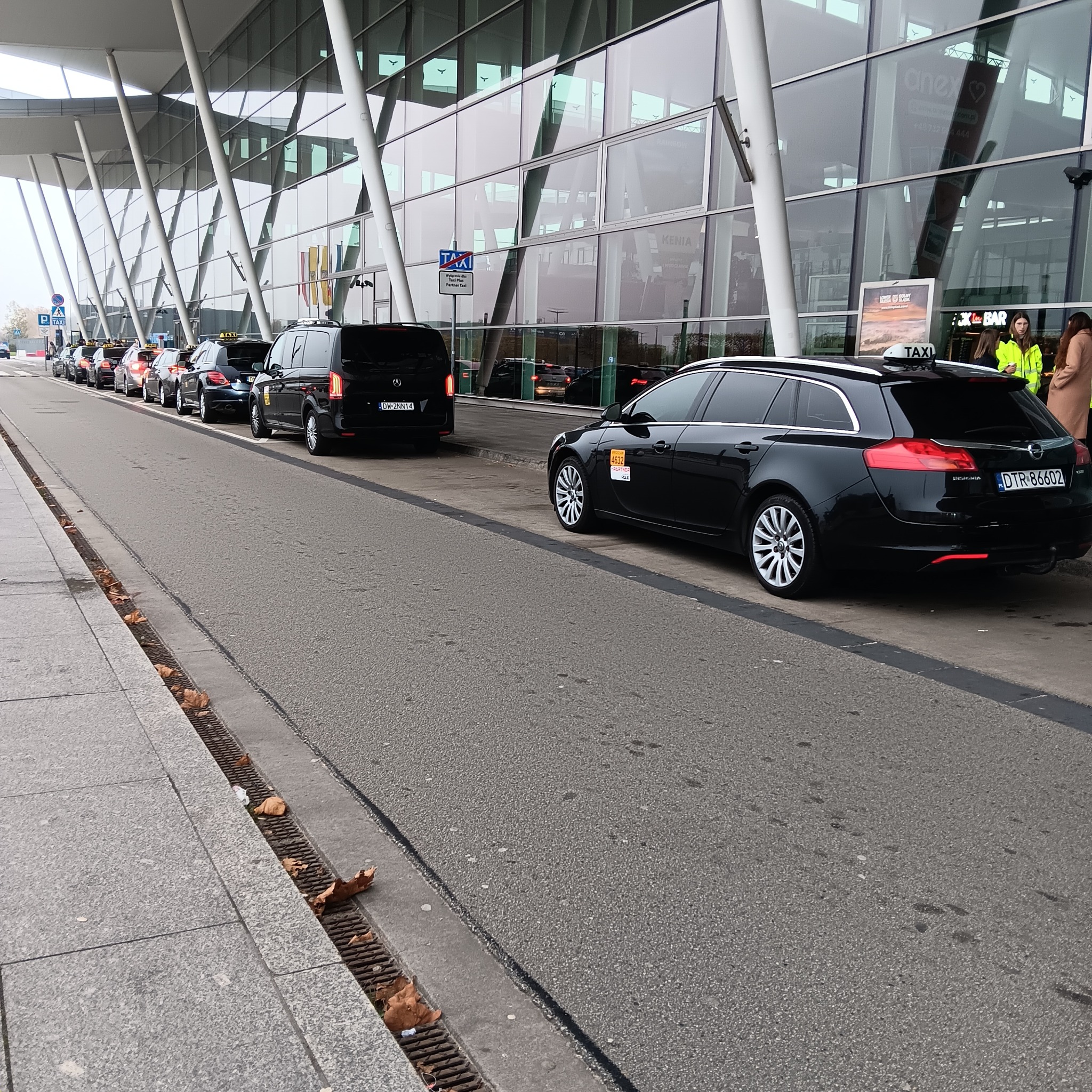 Szereg czarnych taksówek ustawionych przed nowoczesnym budynkiem lotniska z przeszkloną fasadą i białymi kolumnami, widoczne oznaczenia taxi na pojazdach.