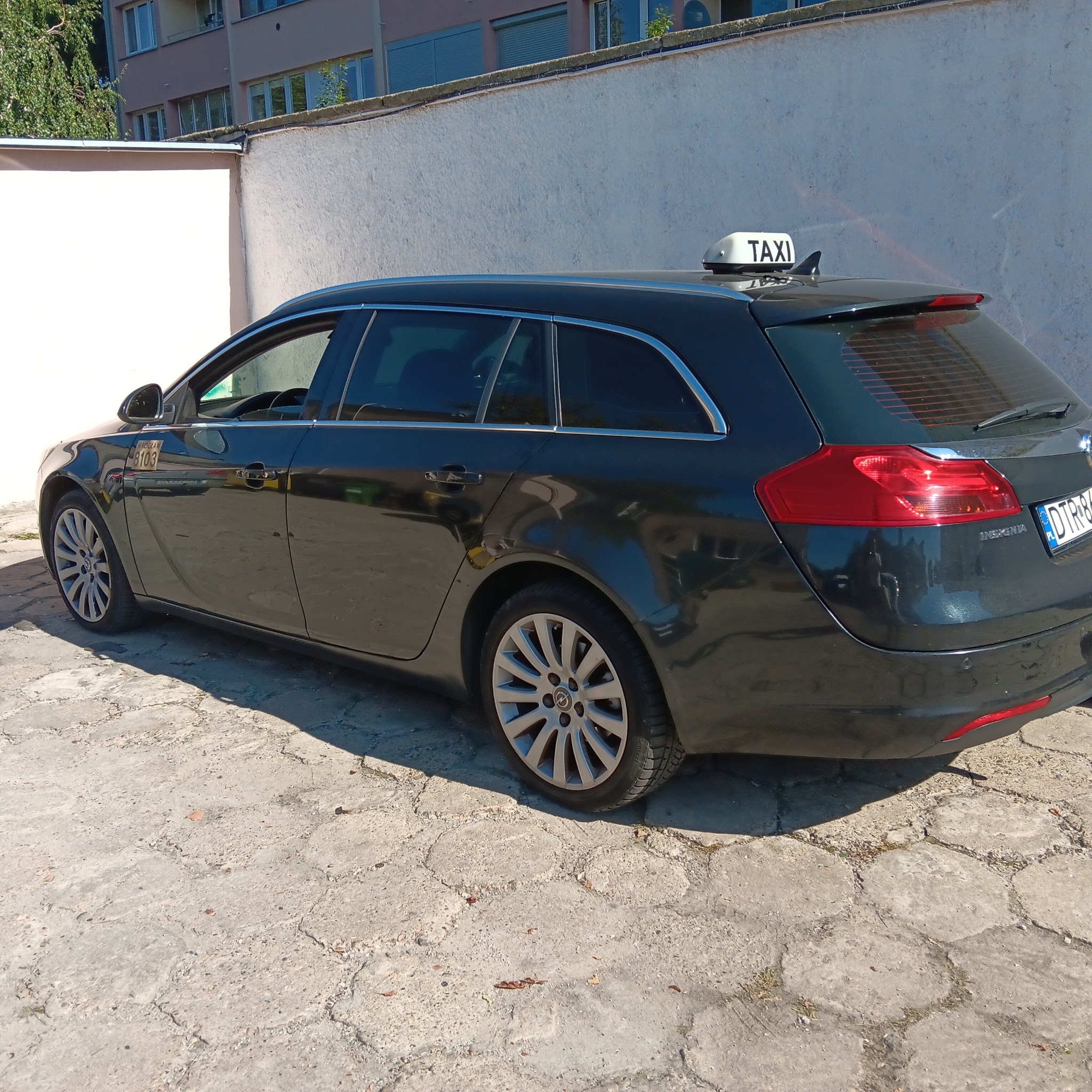 Czarny samochód typu kombi z napisem TAXI na dachu, zaparkowany na betonowym placu, z widoczną naklejką z numerem bocznym i tablicą rejestracyjną.