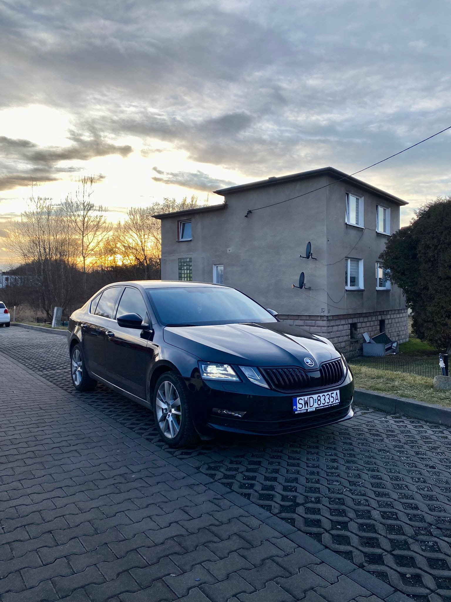 Czarny samochód osobowy marki Skoda zaparkowany na betonowej kostce brukowej przed dwupiętrowym budynkiem mieszkalnym o zachodzie słońca.