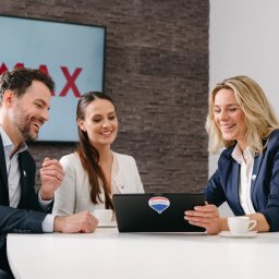 REMAX STEEL - Trzech agentów nieruchomości uśmiechniętych, siedzących przy białym stole z laptopem z naklejką logo firmy, analizujących dane; w tle logo firmy na ekranie i mapa świata.