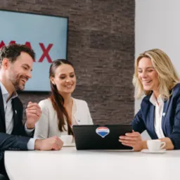 Trzech agentów nieruchomości uśmiechniętych, siedzących przy białym stole z laptopem z naklejką logo firmy, analizujących dane; w tle logo firmy na ekranie i mapa świata.