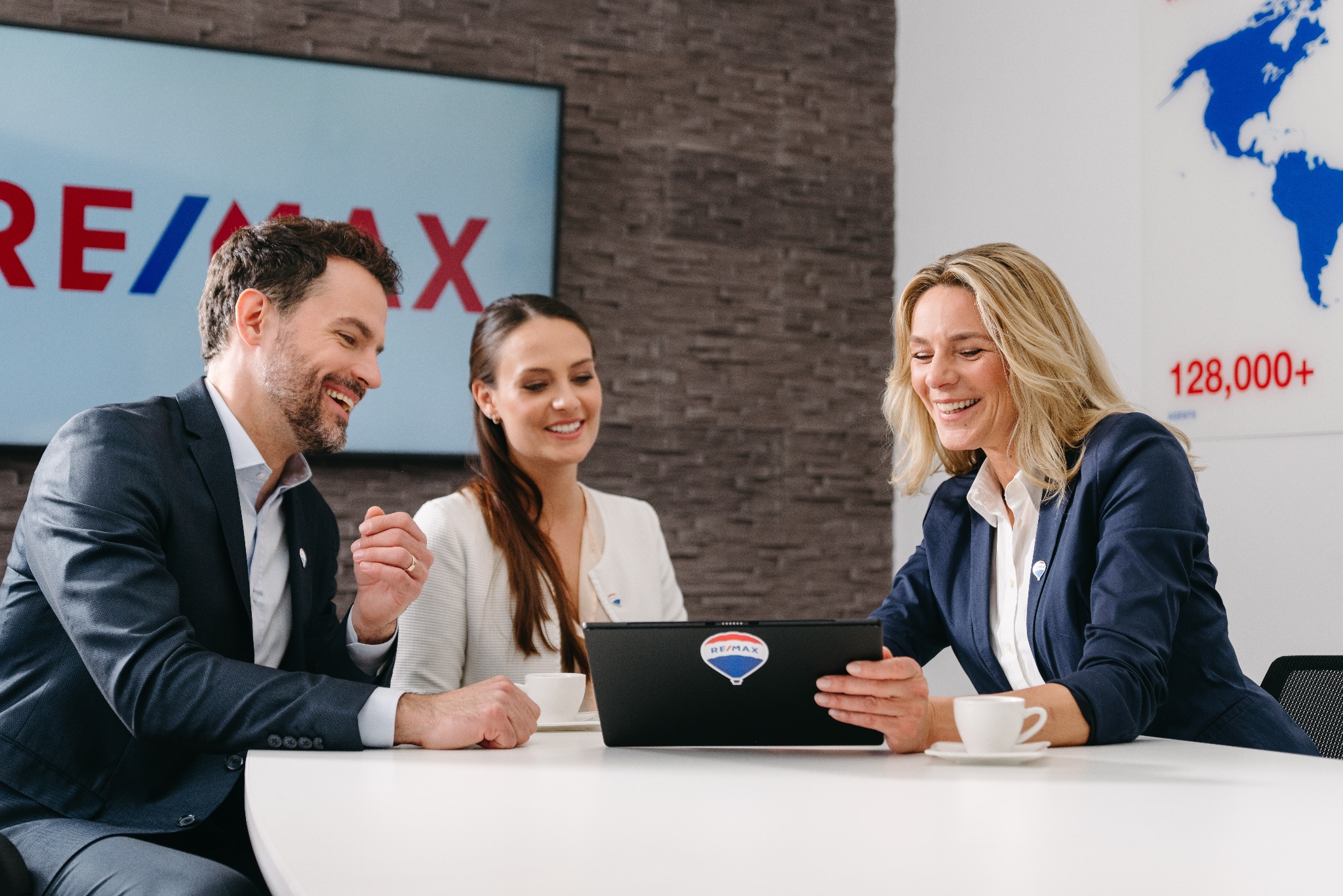 Trzech agentów nieruchomości uśmiechniętych, siedzących przy białym stole z laptopem z naklejką logo firmy, analizujących dane; w tle logo firmy na ekranie i mapa świata.
