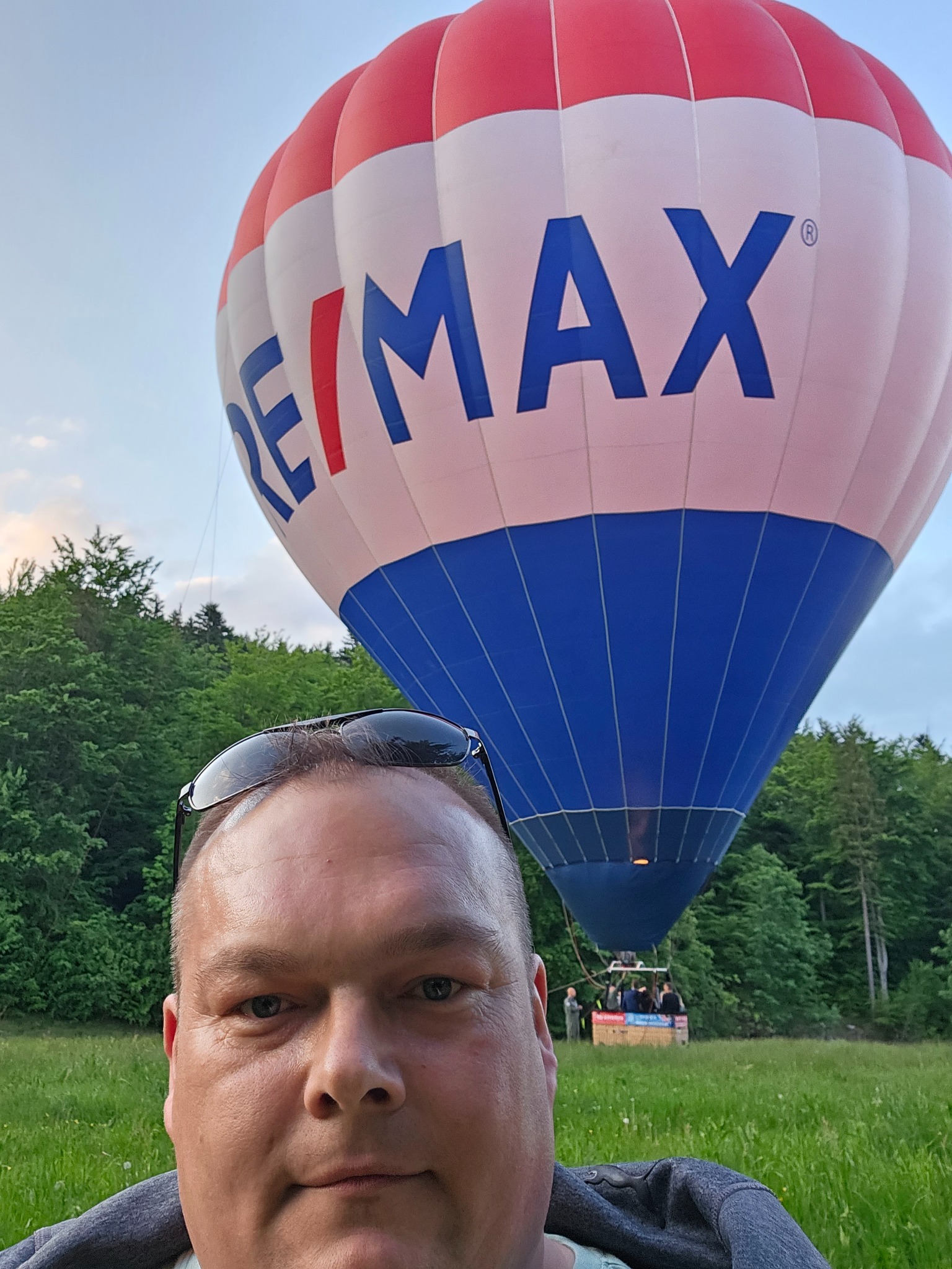 Selfie mężczyzny z okularami na głowie na tle balonu z logo RE/MAX i lasu, w oddali widoczny kosz balonu z pasażerami.