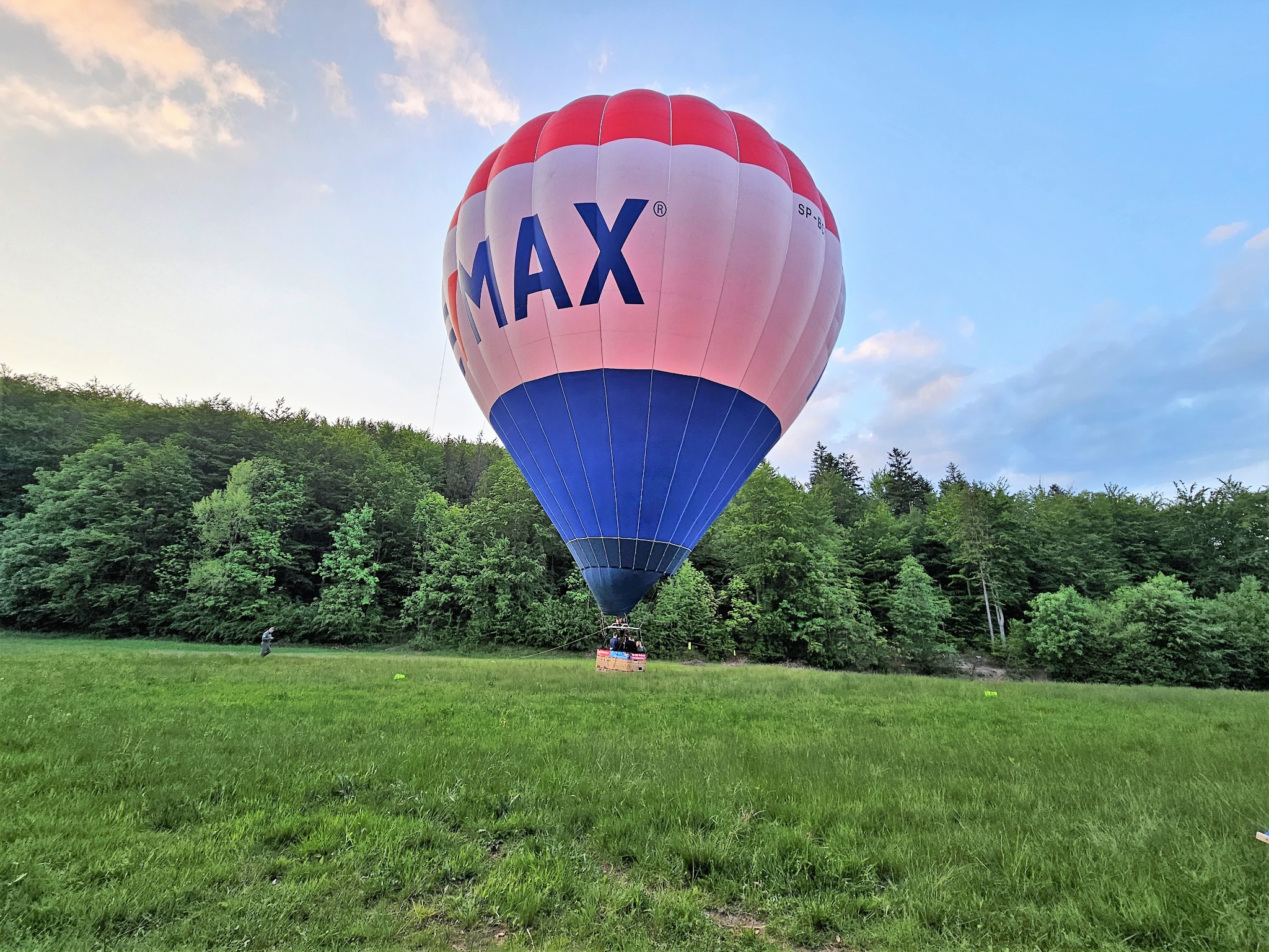 Balon z logo MAX na tle zielonej łąki i lasu, częściowo pochmurne niebo.