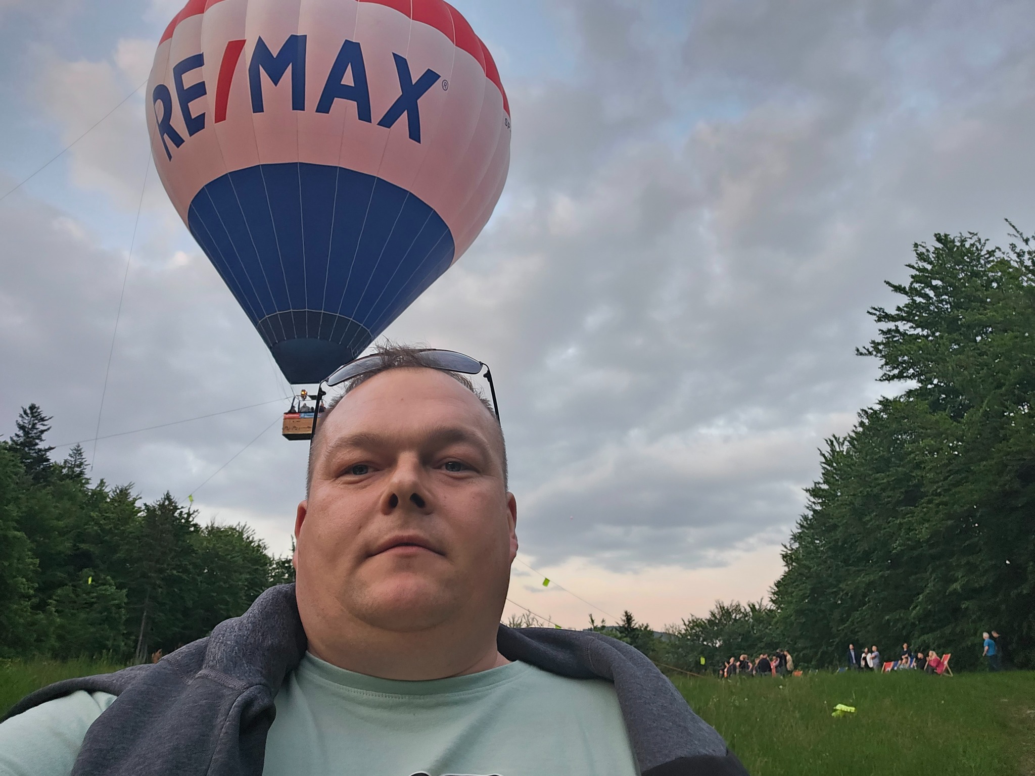 Mężczyzna robiący selfie z balonem RE/MAX w tle, na łące z widzami.