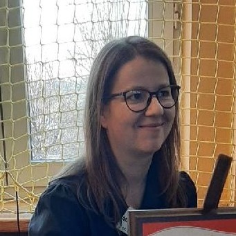 Portret kobiety w okularach, z długimi brązowymi włosami, uśmiechającej się, w tle okno z siatką i fragment plakatu.