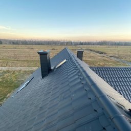 Zbigniew Tarka usługi blacharsko dekarskie - Nowoczesny, grafitowy dach z blachy trapezowej z dwoma kominami na tle rozległego pola i lasu w oddali, sfotografowany z góry w porannym świetle.