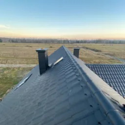 Nowoczesny, grafitowy dach z blachy trapezowej z dwoma kominami na tle rozległego pola i lasu w oddali, sfotografowany z góry w porannym świetle.