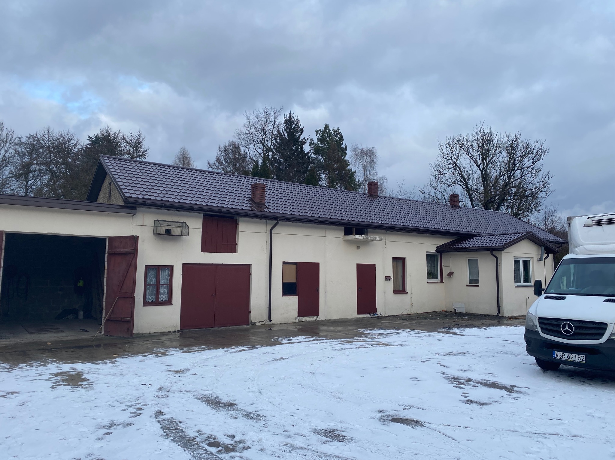 Budynek gospodarczy z nowym, ciemnobrązowym dachem z blachodachówki, widoczne kominy i rynny, na pierwszym planie częściowo ośnieżony plac i samochód dostawczy.
