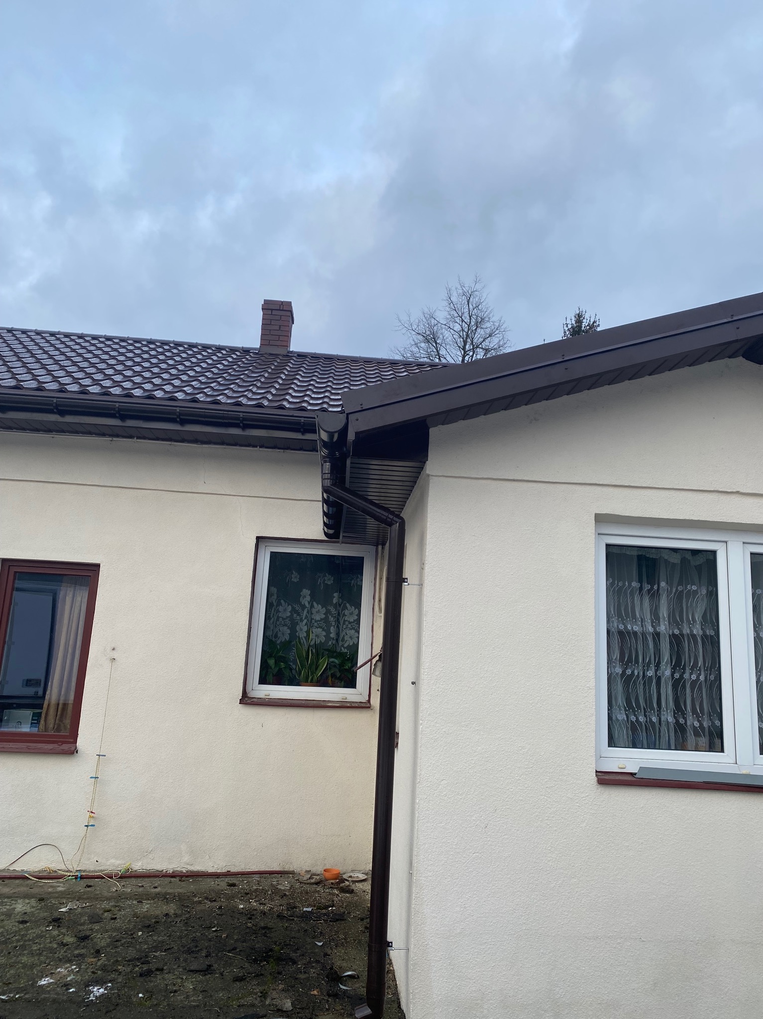 Fragment domu z brązowym dachem z dachówki i rynnami, widoczne okna i komin na tle pochmurnego nieba.