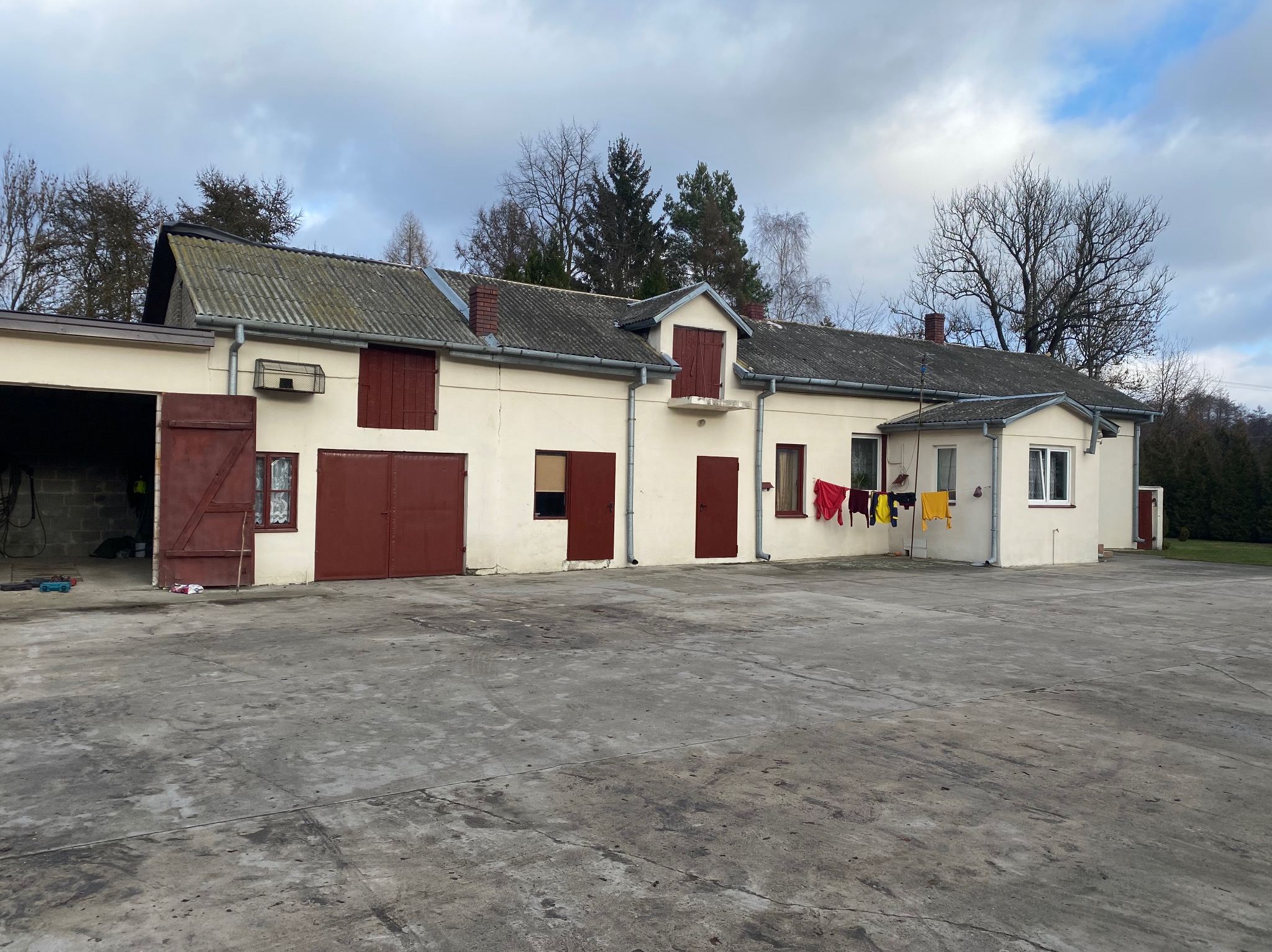 Budynek gospodarczy z szarym dachem z eternitu, czerwonymi okiennicami i rynnami, na podwórzu suszy się pranie.