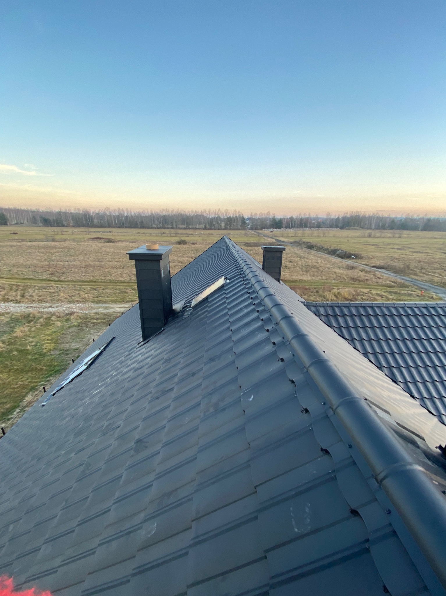 Nowoczesny, grafitowy dach z blachy trapezowej z dwoma kominami na tle rozległego pola i lasu w oddali, sfotografowany z góry w porannym świetle.