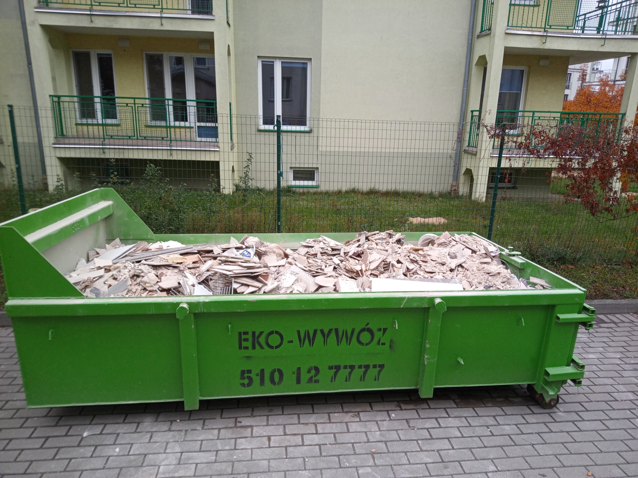 Kontener EKO-WYWÓZ wypełniony gruzem budowlanym na tle bloku mieszkalnego z zielonym ogrodzeniem. Widoczny numer telefonu.