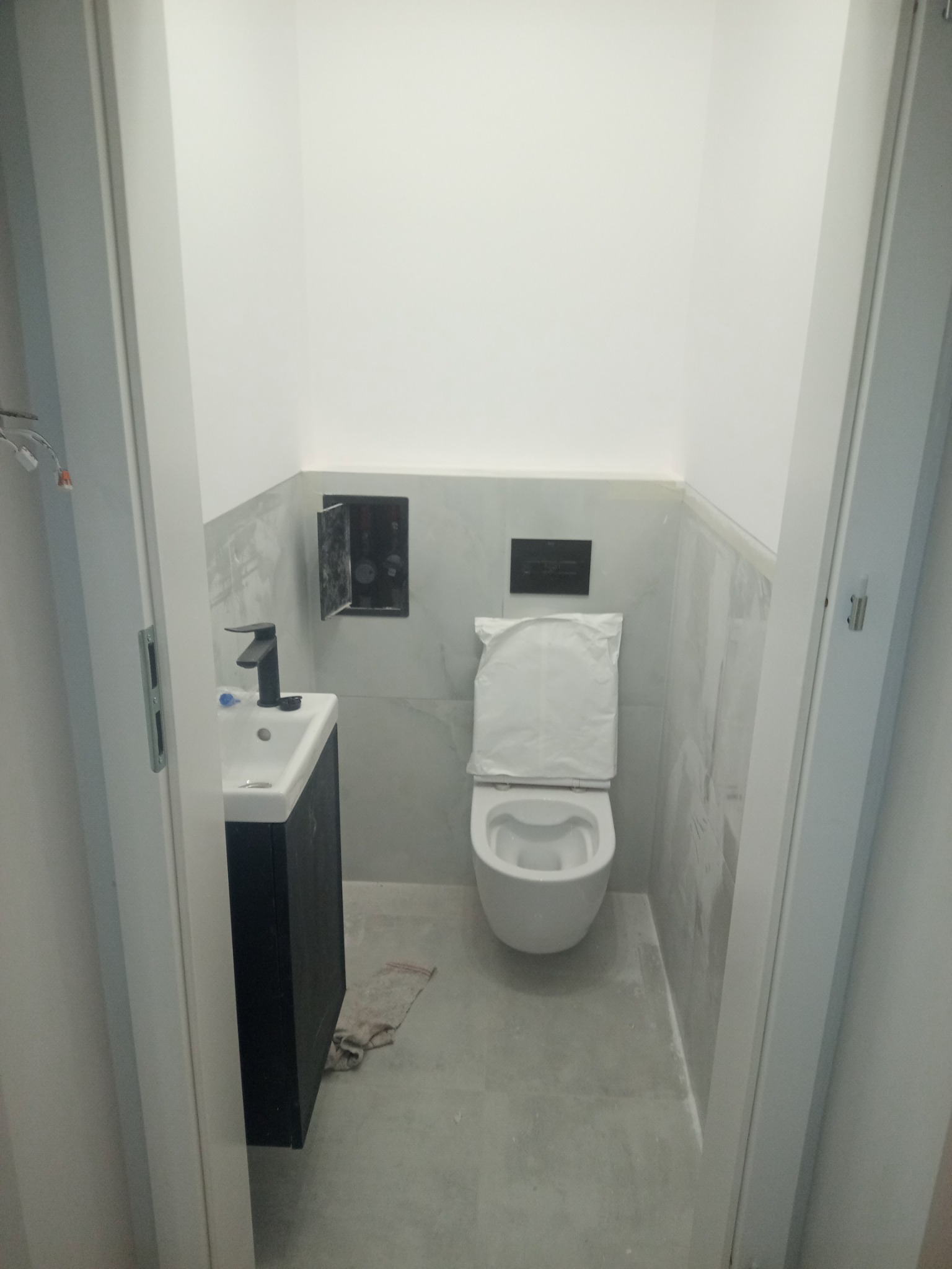 Nowoczesna łazienka w trakcie wykańczania: biała toaleta z osłoną, umywalka z czarną szafką i szare płytki na ścianach. Widok zza otwartych drzwi.