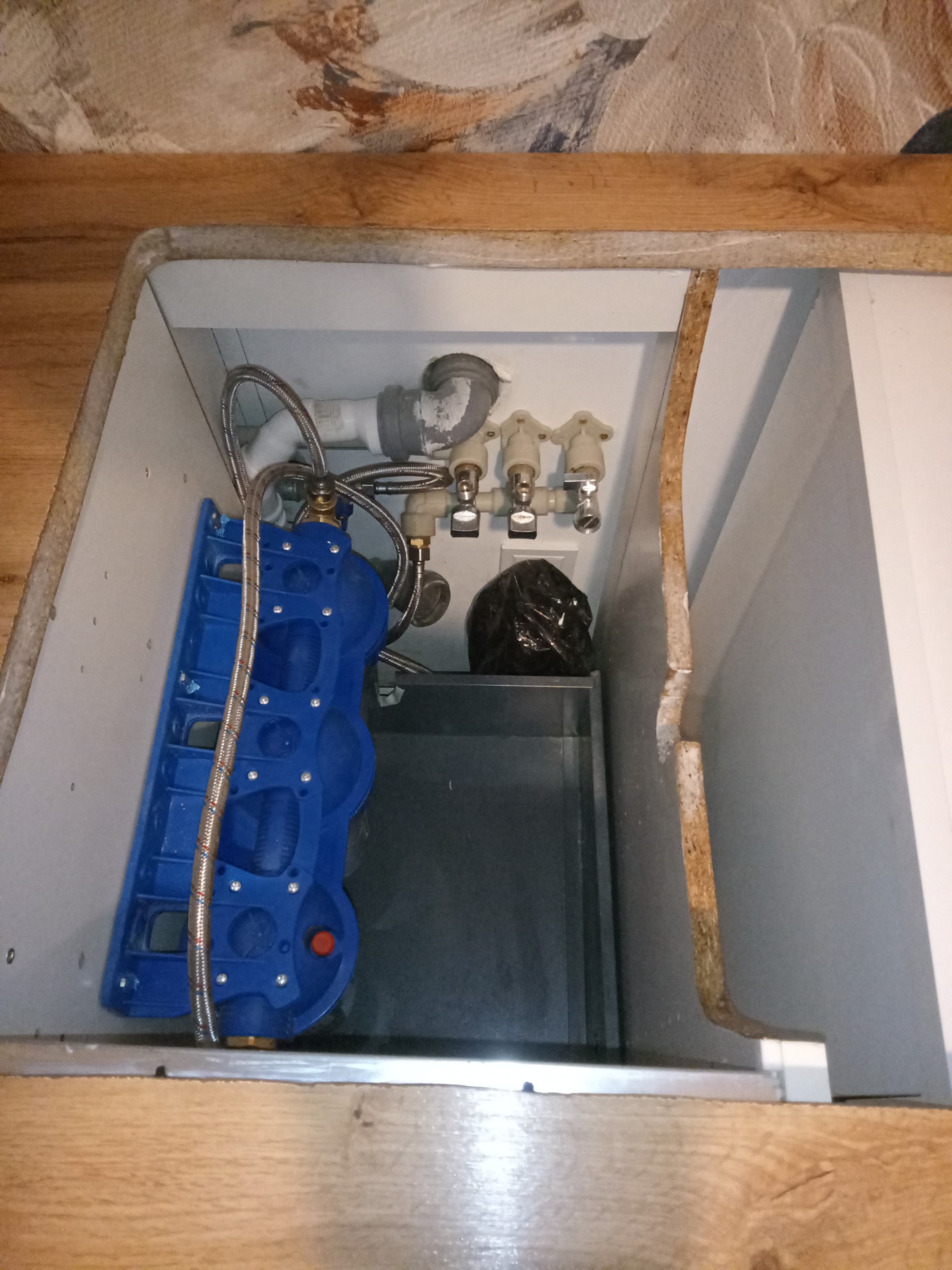 Wnętrze szafki pod zlewem z widoczną instalacją hydrauliczną, w tym zawory, węże w oplocie i syfon. Widoczny także filtr wody i odpływ kanalizacyjny.