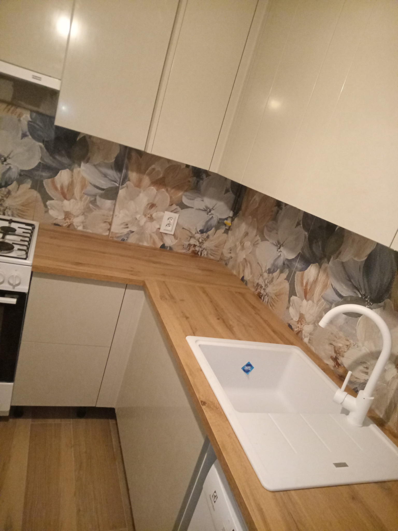 Jasna kuchnia po remoncie: białe szafki, drewniany blat, kwiatowe płytki na ścianie i biały zlew z baterią. Widoczny fragment kuchenki i podłogi.