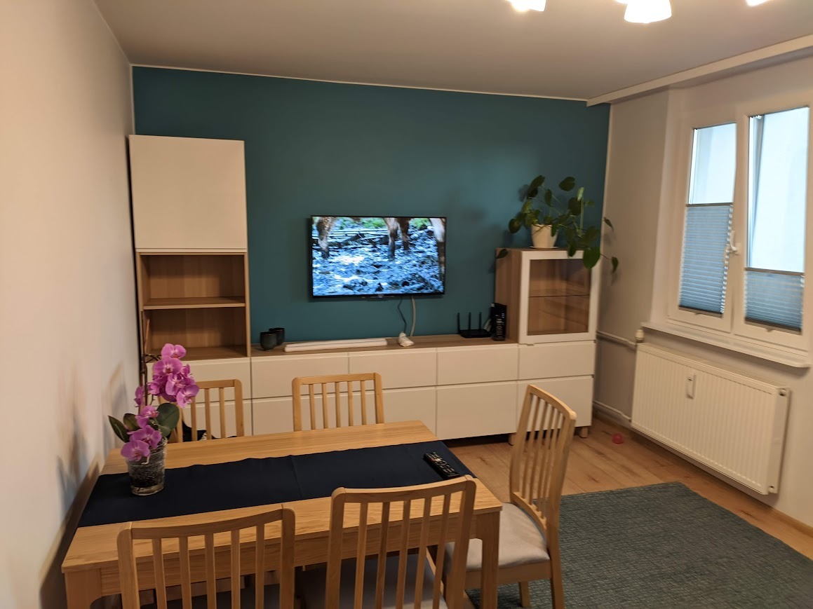 Jasne drewniane krzesła i stół z czarnym bieżnikiem w nowoczesnym salonie z turkusową ścianą, białymi meblami modułowymi i telewizorem.