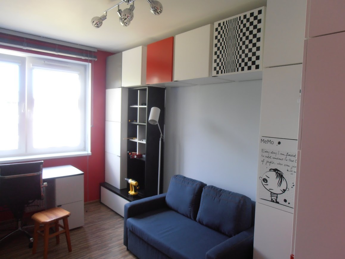 Pokój z zabudową meblową: szafki wiszące w kolorach białym, pomarańczowym i szachownicy, regał z półkami, niebieska sofa, biurko, krzesło i lampa podłogowa. Na regale widoczna żółta wkrętarka.