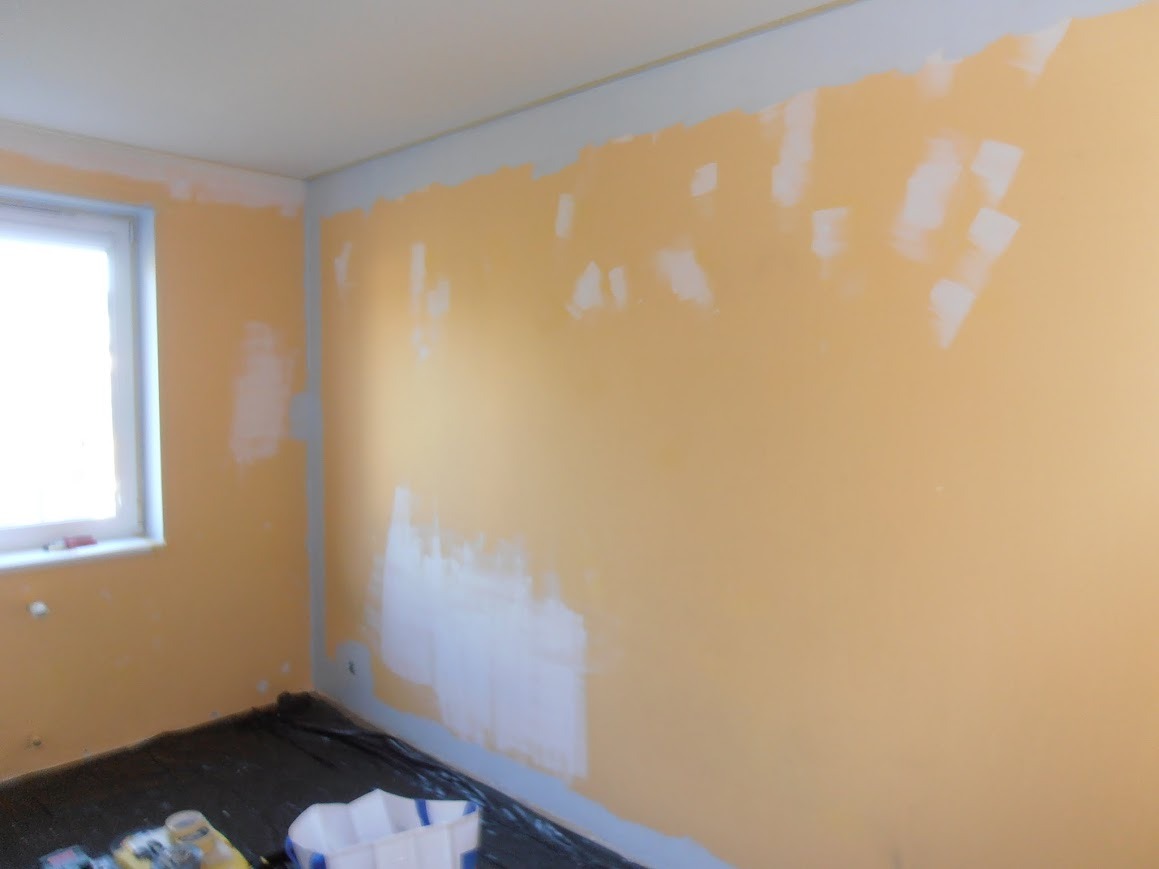 Pokój w trakcie malowania na żółto, z białymi plamami szpachli na ścianach, zabezpieczona podłoga, widoczne wiadro i narzędzia malarskie.
