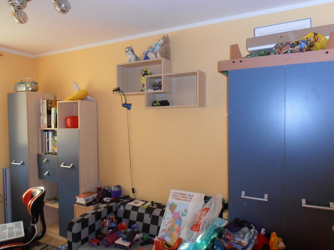 Pokój dziecięcy z niebieską szafą, regałami z książkami i zabawkami, oraz półkami na ścianie z pluszakami i robotem.