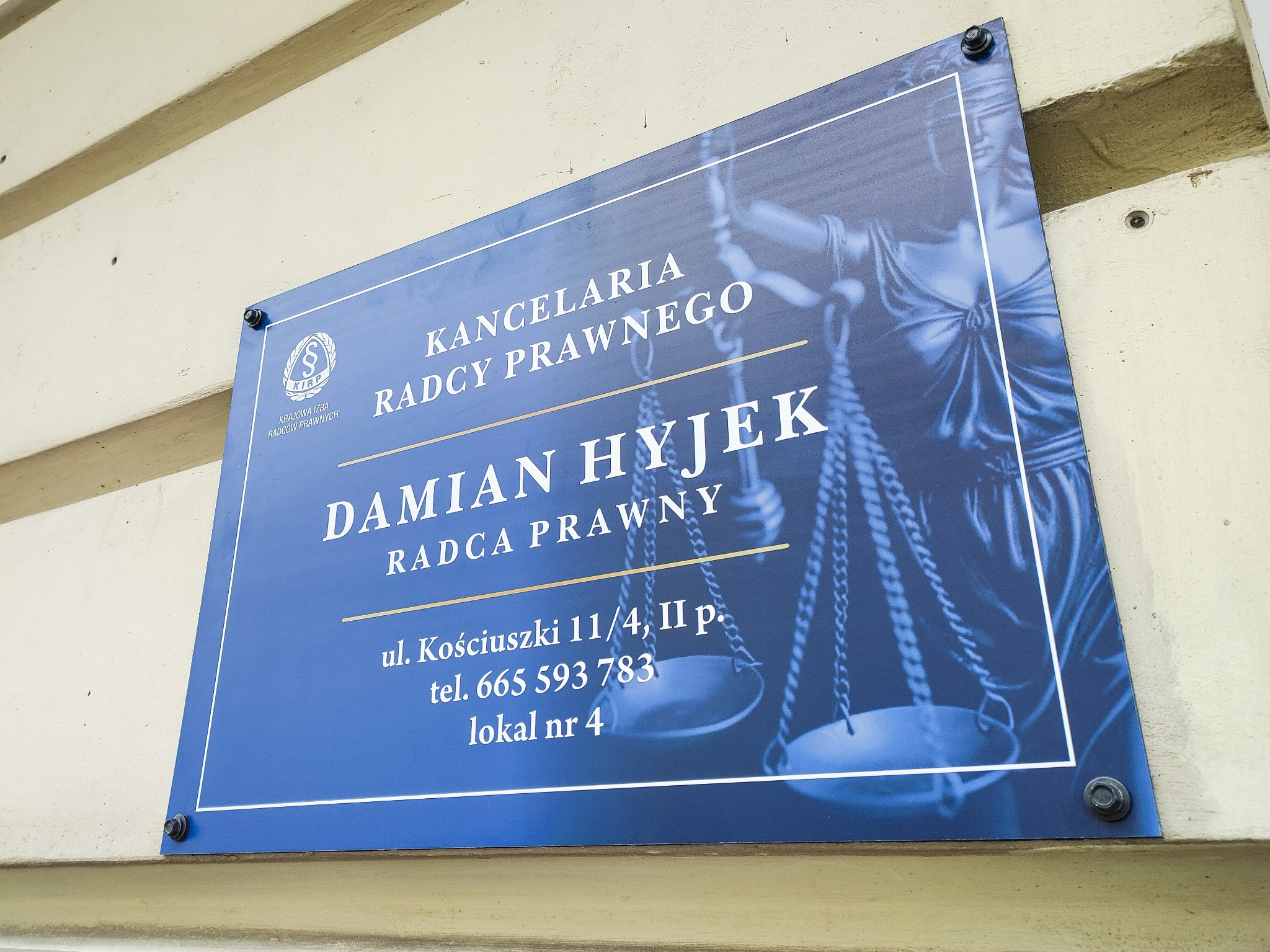 Szyld Kancelarii Radcy Prawnego Damiana Hyjek z adresem, numerem telefonu i symbolem Temidy, przymocowany do ściany budynku.