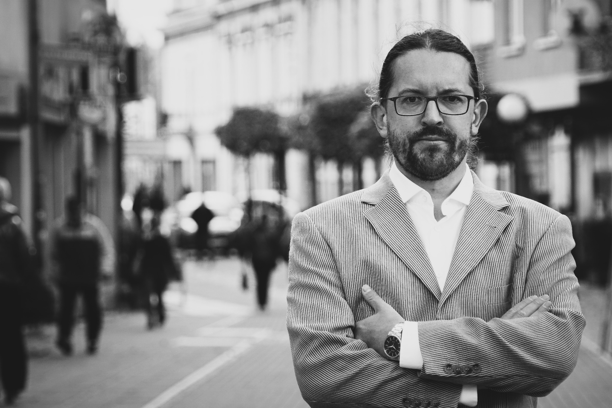 Czarno-białe portretowe zdjęcie mężczyzny w okularach i marynarce w paski na tle miejskiej ulicy z przechodniami.