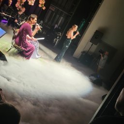 Grzegorz Baster Eventy - Dwie wokalistki na scenie w dymie, jedna w bordowej sukni siedzi na ławce, druga w zielonej sukni wieczorowej stoi, w tle zespół muzyczny i kamera.