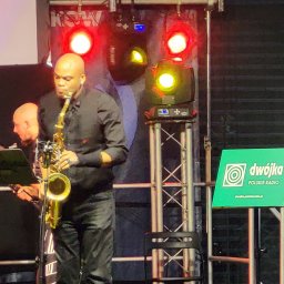 Grzegorz Baster Eventy - Saksofonista w czarnej koszuli gra na saksofonie podczas koncertu na scenie z oświetleniem i logo Dwójki Polskie Radio w tle.