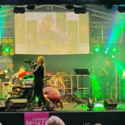 Scena plenerowa podczas festiwalu jazzowego, wokalistka w czarnej sukni śpiewa do mikrofonu, w tle muzycy grają na instrumentach, oświetlenie sceniczne w kolorze zielonym, na ekranie wyświetlają...