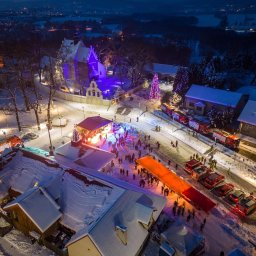 Grzegorz Baster Eventy - Widok z lotu ptaka na zimowy wieczór z oświetloną sceną, kościołem w fioletowym świetle i wozami strażackimi w tle.